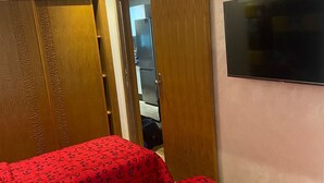 Ein Schlafzimmer mit einem Bett, das mit einer roten blumigen Tagesdecke bezogen ist, Holzschränken und einem an der Wand befestigten Flachbildfernseher.