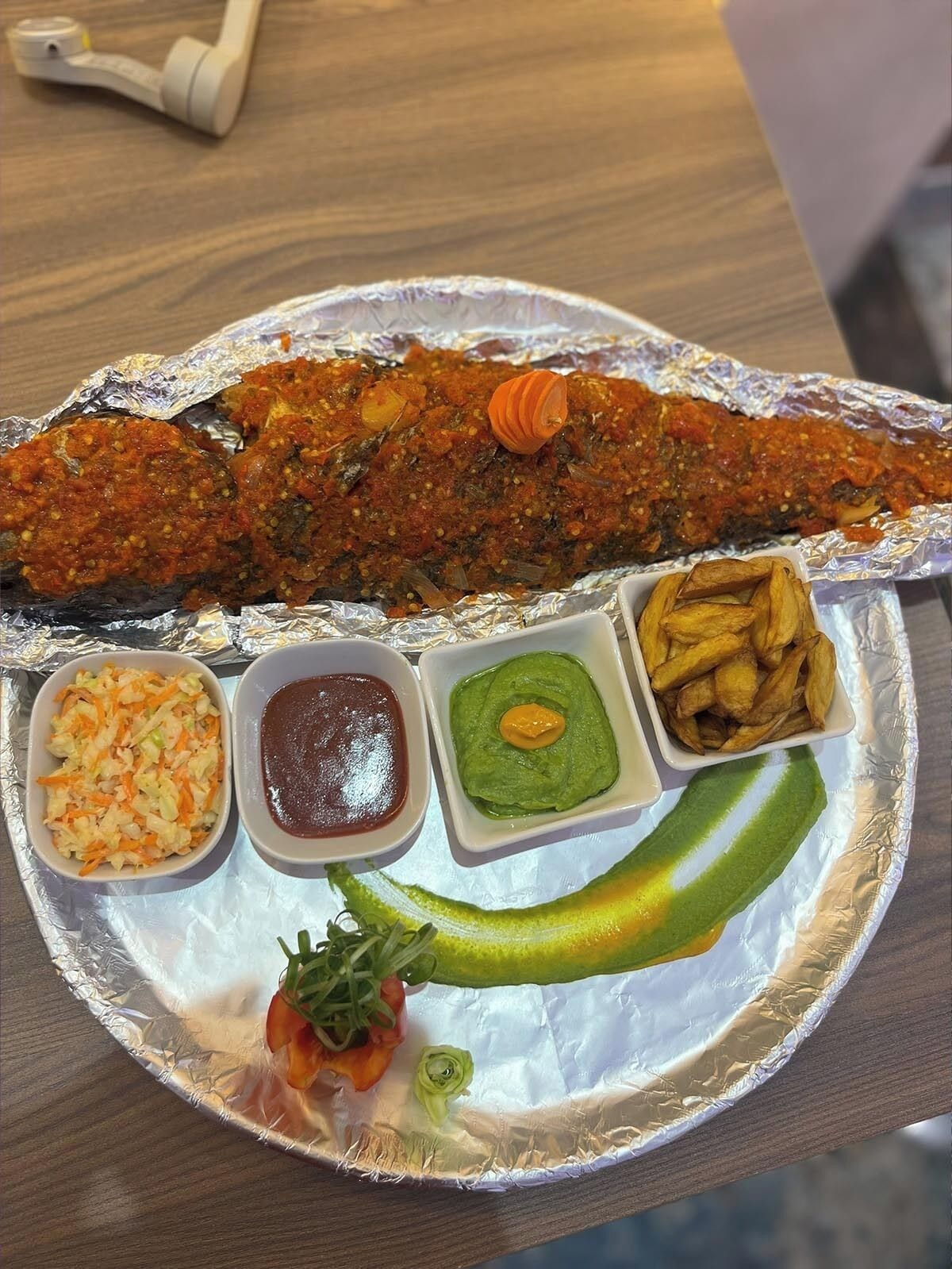 A meal with a whole fish, coleslaw, green sauce, and fries on a foil-lined tray.