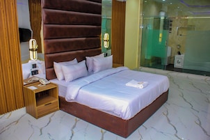 Quarto de hotel moderno com uma cama grande, cabeceira de madeira, mesas de cabeceira e box de chuveiro em vidro.