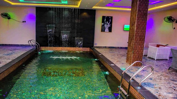 A swimming pool with a waterfall feature, surrounded by lounge chairs and a brick column. - Soulmate Hotels & Suites Ojodu (Lagos)