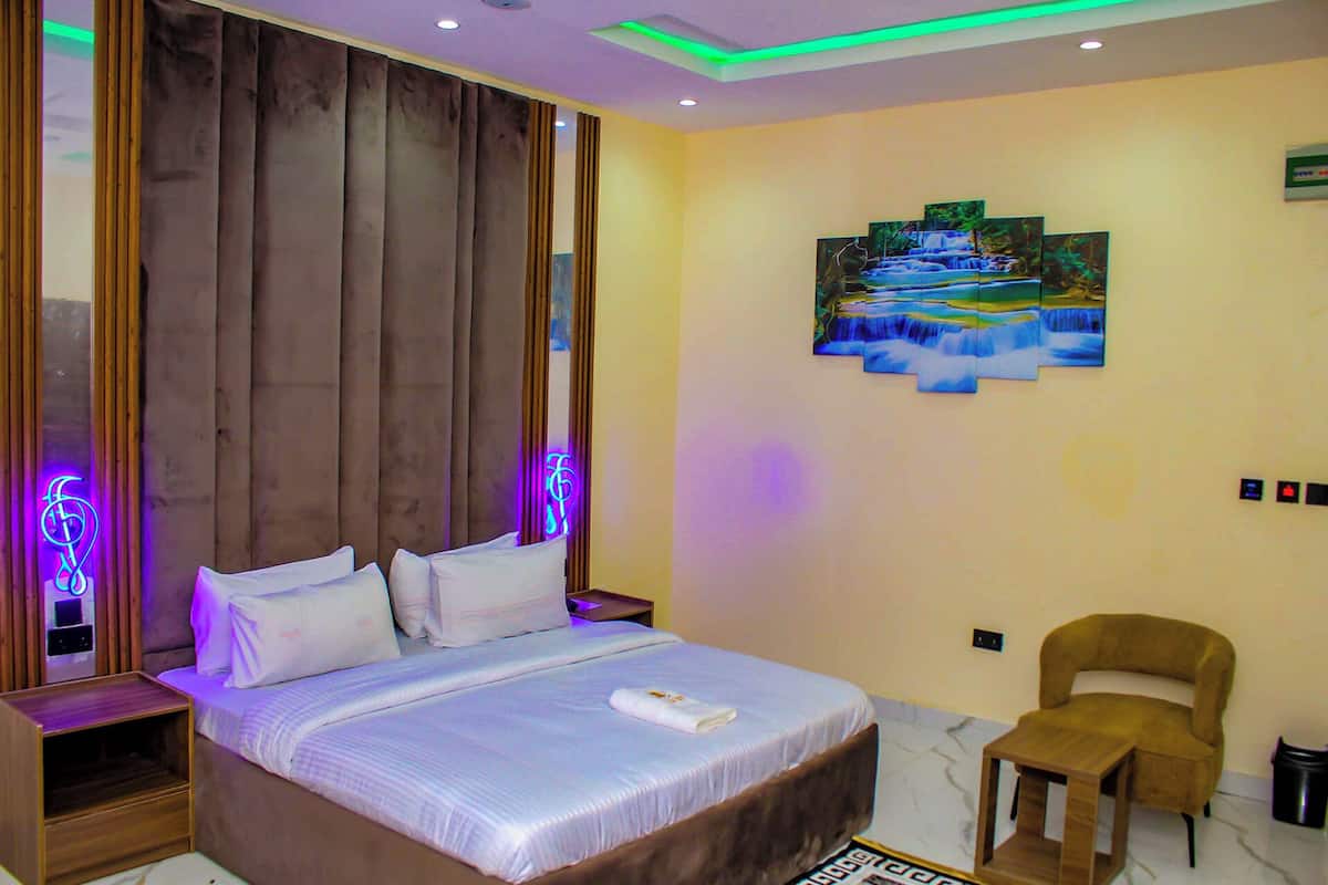 A hotel room with a bed, a chair, a painting, and a neon sign.