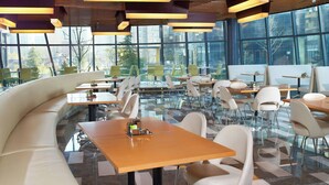 A modern dining area with a mix of seating options including a curved bench and individual tables, large windows offering a view of the city, and a ceiling with wooden slats and hanging lights. - Aloft Haidian Hotel Beijing (Beijing)