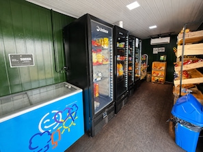A refrigerated display area with shelves stocked with various food items, a blue cooler with a colorful logo, and a trash bin. - Hosts BR - Viva Praia do Futuro (Fortaleza)