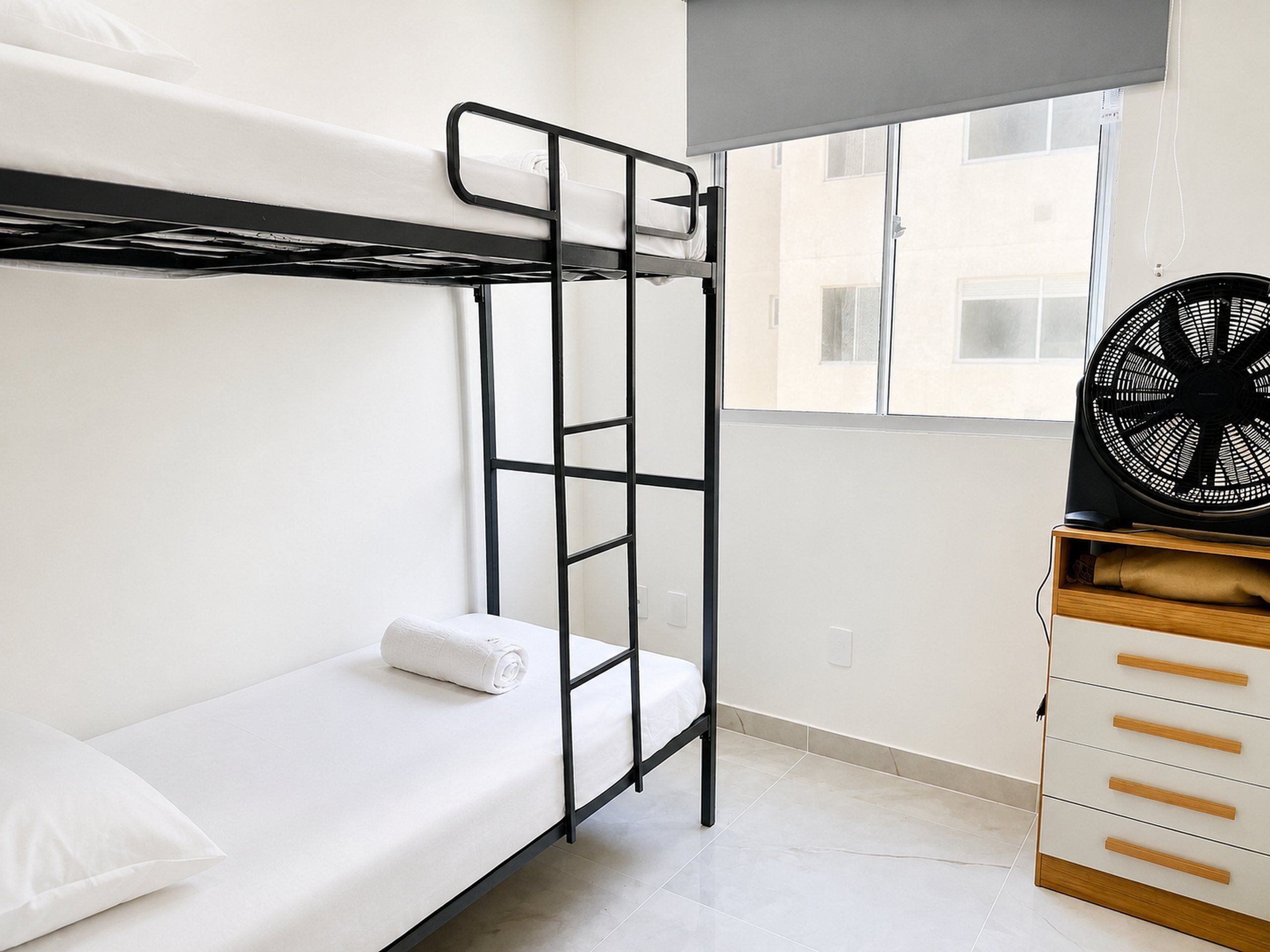 Uma cama de beliche com escada, um ventilador e um armário de madeira num quarto com janela.