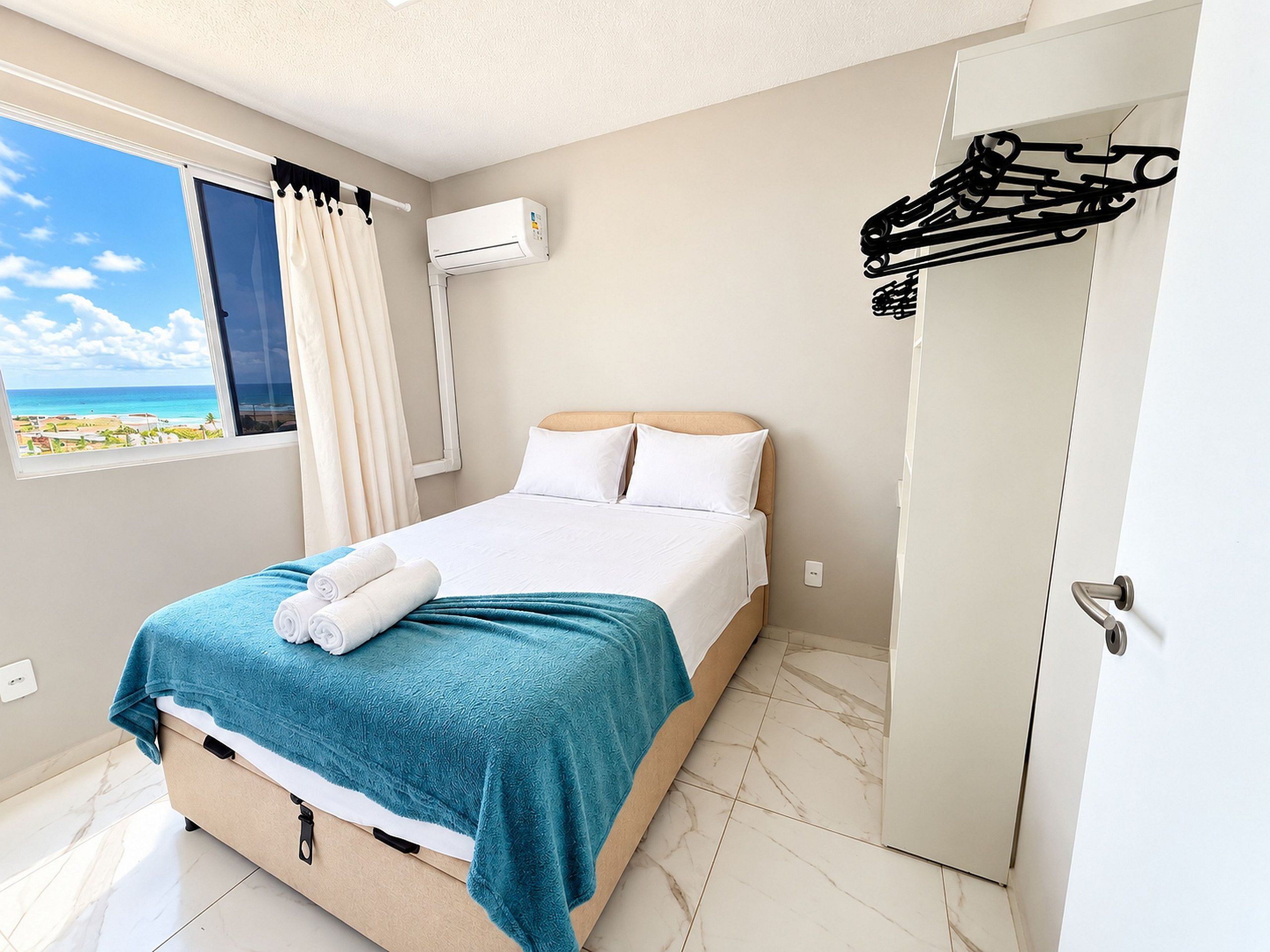 Una habitación de hotel con cama, ventana con cortinas, equipo de aire acondicionado y puerta con perchero.