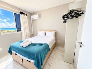 A hotel room with a bed, a window with curtains, an air conditioning unit, and a door with a coat rack.