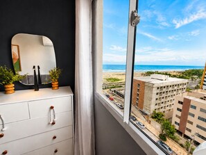 A room with a dresser, a mirror, and a view of a beach and buildings. - Hosts BR - Viva Praia do Futuro (Fortaleza)