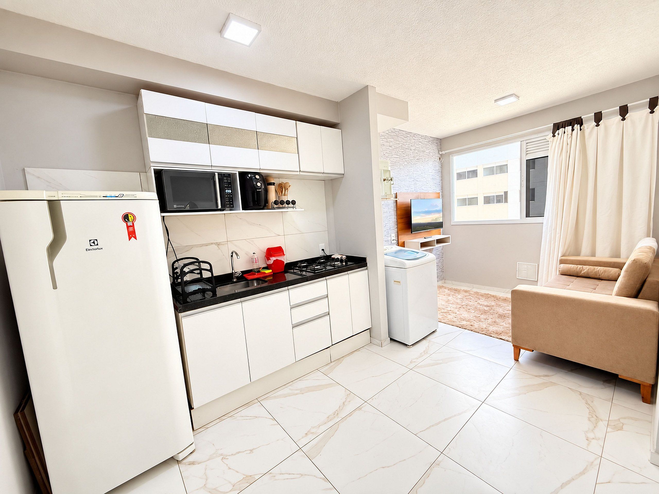 A compact kitchen with a refrigerator, microwave, and stove, a washing machine, and a sofa in a living area with a view of a building outside.