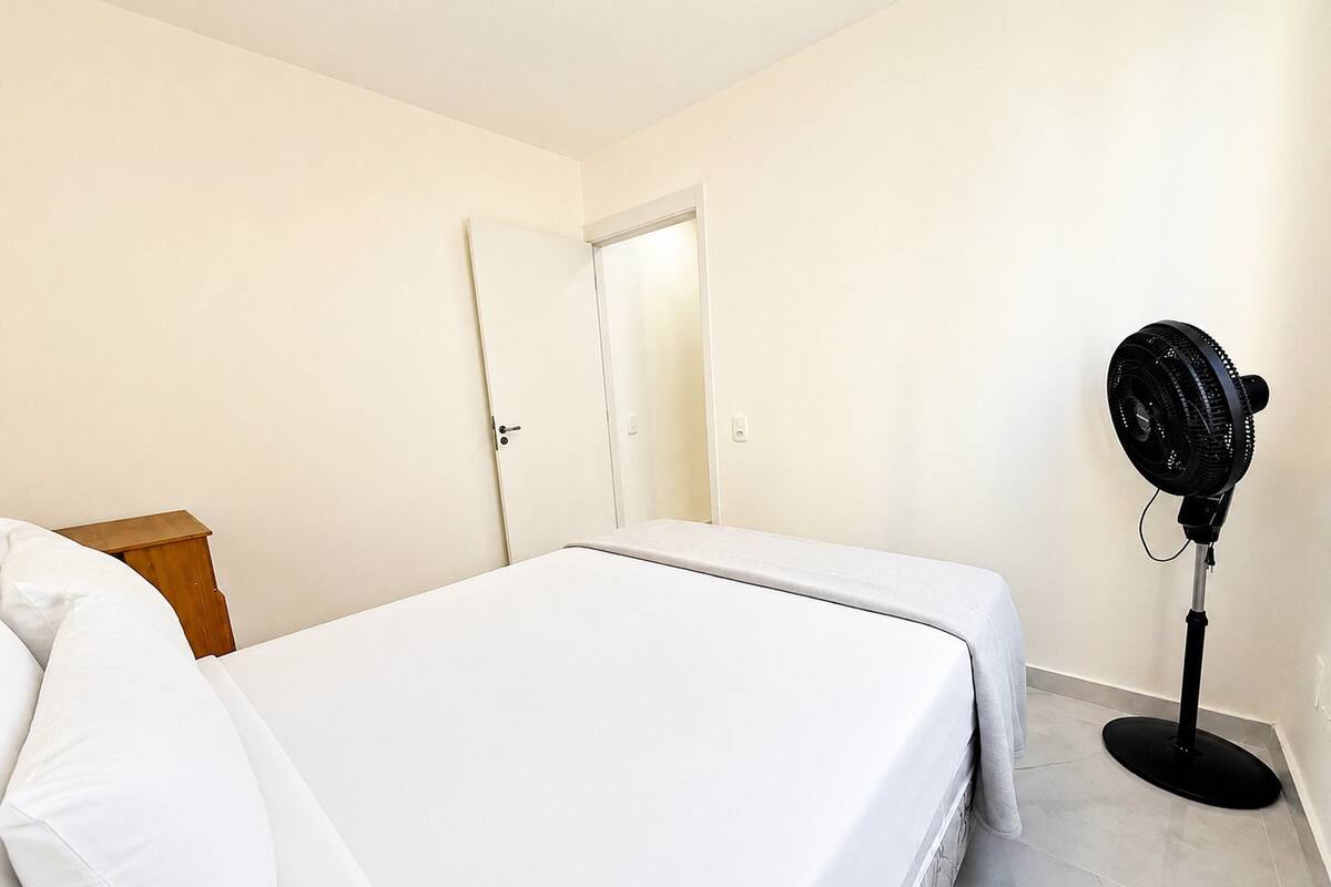 Un dormitorio con cama, mesita de noche, puerta y ventilador de pie.