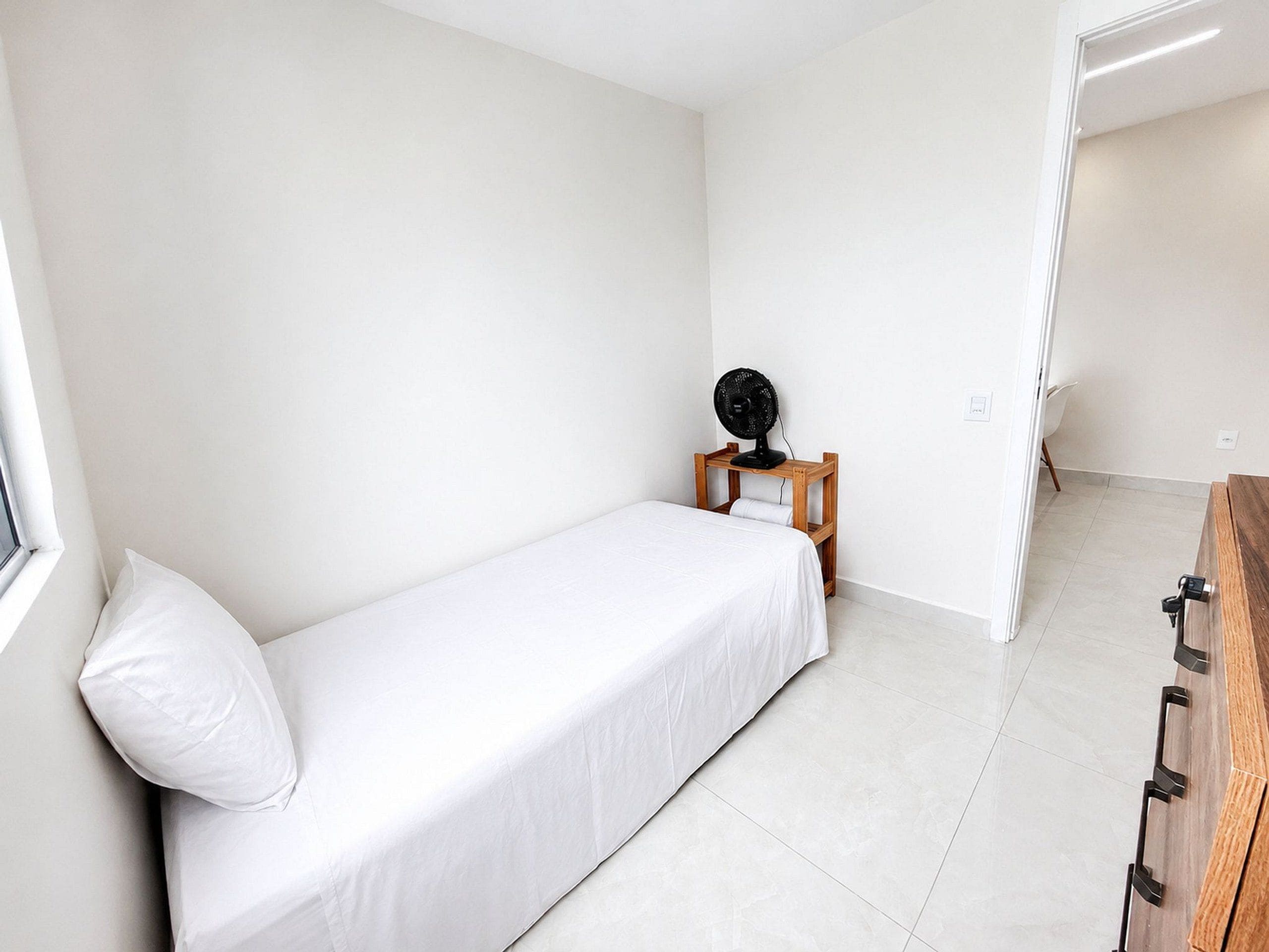 A small, single-bed room with a wooden bedside table, a fan, and a window.
