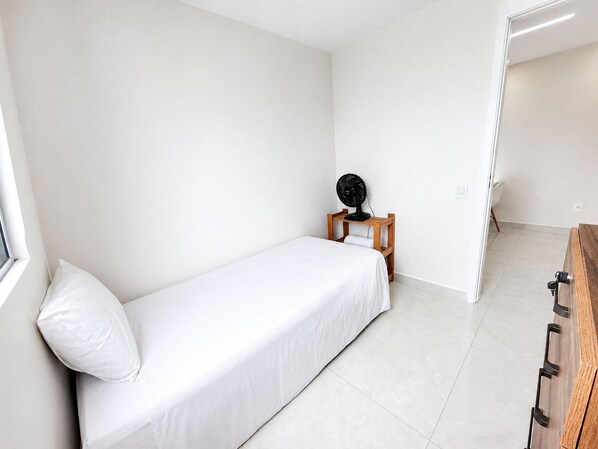A small, single-bed room with a wooden bedside table, a fan, and a window. - Hosts BR - Viva Praia do Futuro (Fortaleza)