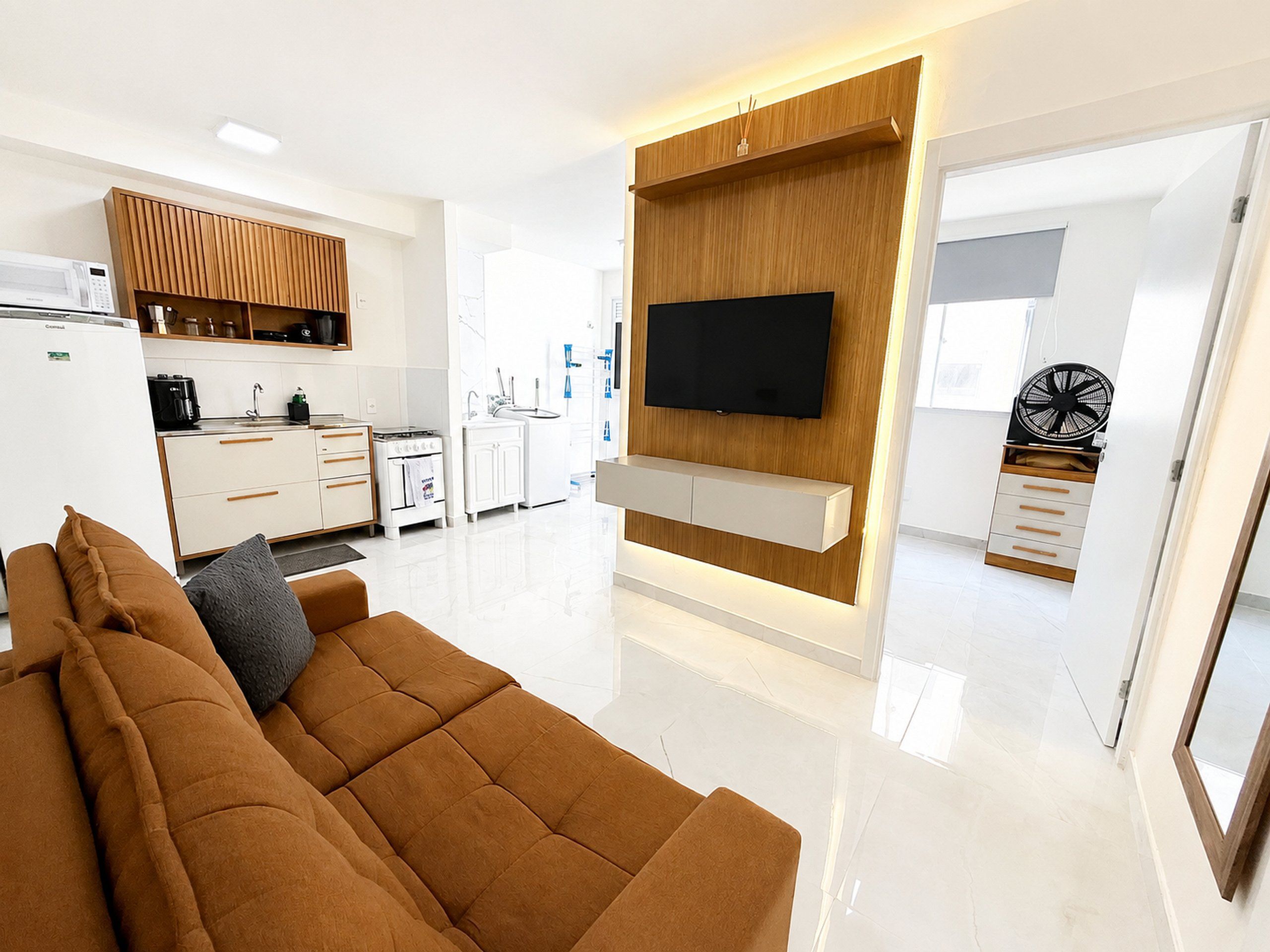 Un salón moderno con un sofá marrón, un televisor de pantalla plana montado en una pared de madera, una zona de cocina con electrodomésticos blancos y un ventilador en la esquina.