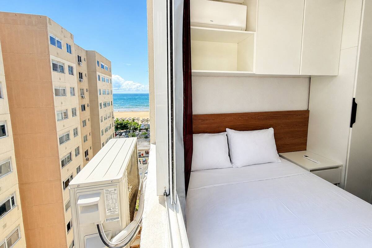 Habitación de hotel con cama, ropa de cama blanca, cabecero de madera y vistas a la playa y edificios.