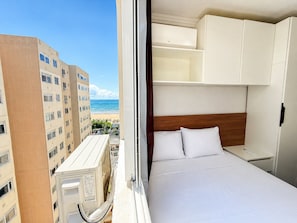 Habitació d'hotel amb un llit, roba de llit blanca, un capçal de llit de fusta i vista a la platja i els edificis.