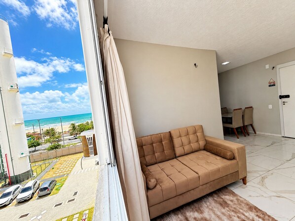 A living room with a beige sofa, a dining area with chairs, a white door, and a view of the beach and ocean through a window.