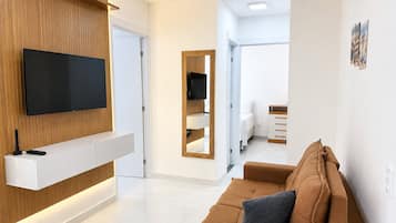 A modern living room with a flat-screen TV, a wooden accent wall, a brown sofa, and a hallway leading to a bedroom.