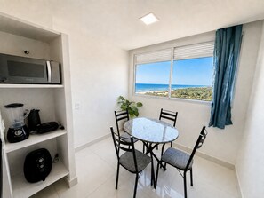 A small dining area with a table and chairs, a microwave above the table, and a window with a view of the beach.