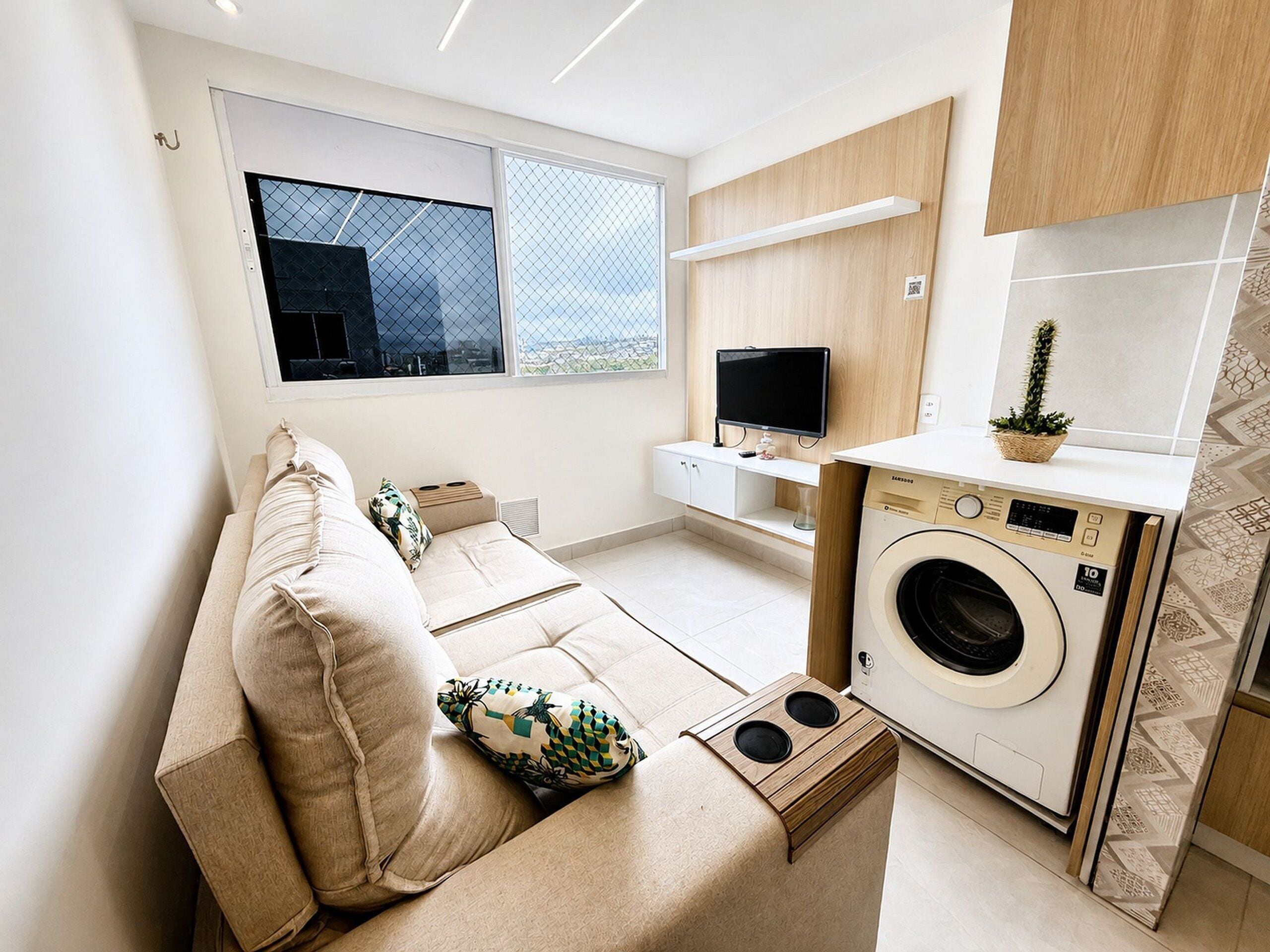 A compact living space with a sofa, a washing machine, a television, and a window with a view.