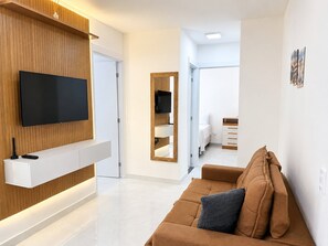 A modern living room with a flat-screen TV, a wooden accent wall, a brown sofa, and a hallway leading to a bedroom. - Hosts BR - Viva Praia do Futuro (Fortaleza)