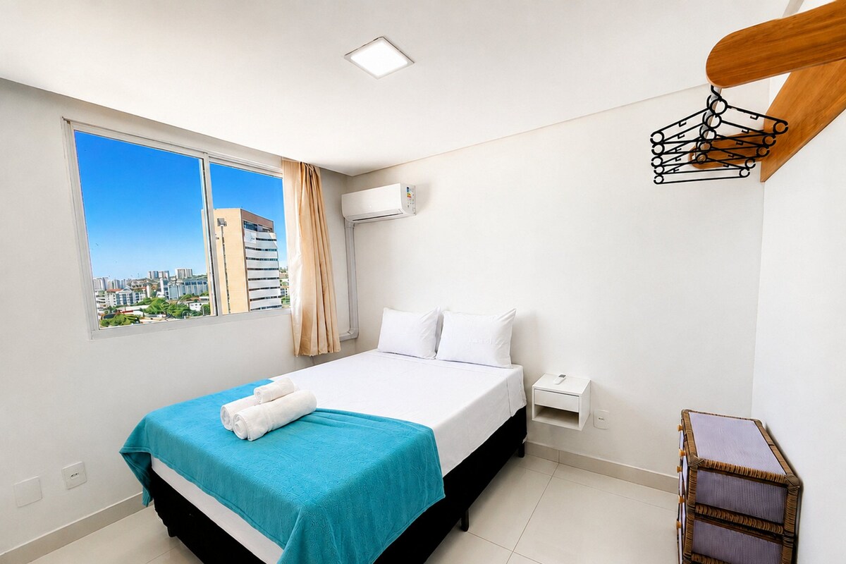 Una camera d'albergo con un letto, un comodino, un condizionatore d'aria, una finestra con vista sulla città e una sedia di legno.