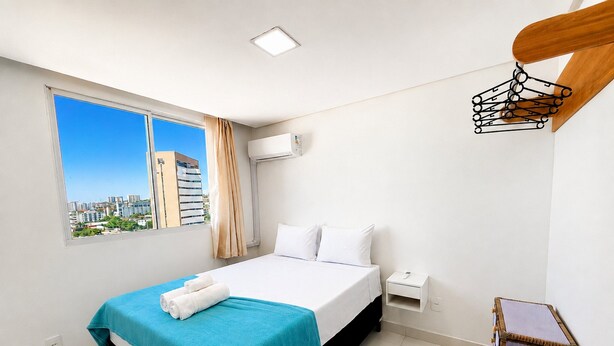 A hotel room with a bed, a nightstand, an air conditioning unit, a window with a city view, and a wooden chair.