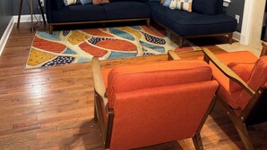 Una sala de estar con un sofá azul oscuro, sillones naranja, un tapete colorido y una lámpara de pie.