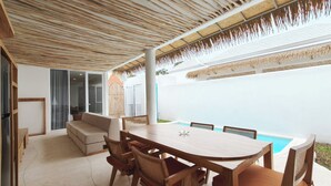 Un espacio de comedor al aire libre cubierto, con piscina, mobiliario de madera y una pared exterior blanca.