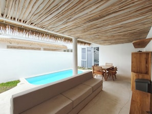 Un área de piscina moderna al aire libre con techo de madera, un banco integrado y una mesa de comedor con sillas.