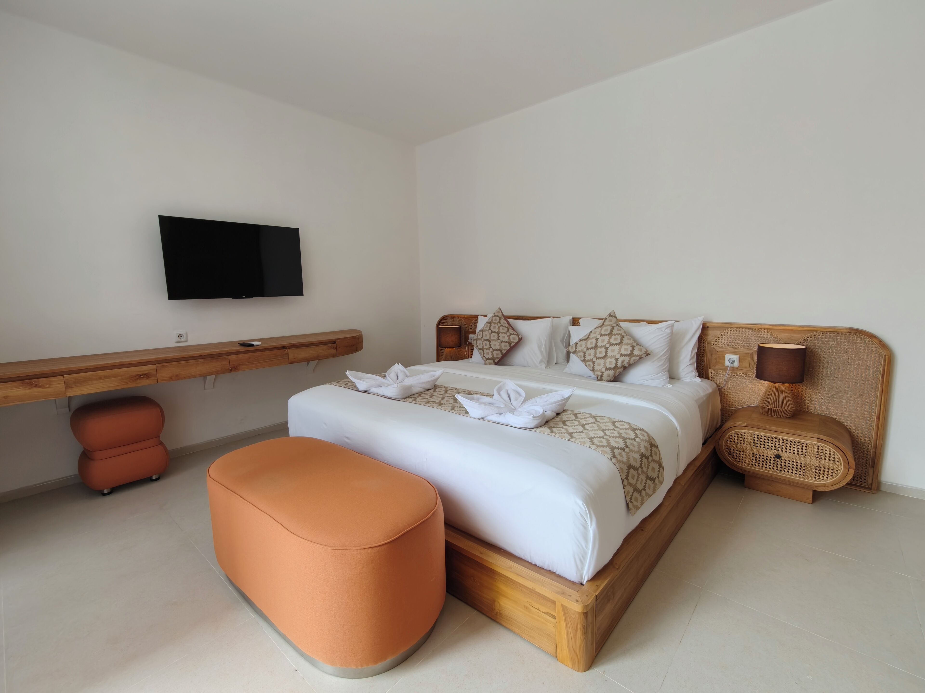 A neatly arranged bedroom with a bed, a wooden bedside table, a television mounted on the wall, and a small orange ottoman.