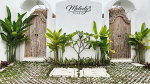 Una villa blanca con un letrero que dice "Melody's Villa" y dos puertas de madera con intrincados grabados.