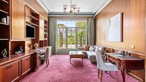 A spacious room with a large wooden desk, a comfortable sofa, and a television. There is a large window with a view of greenery outside.