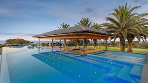 A large infinity pool with a wooden gazebo, palm trees, and a view of the ocean.