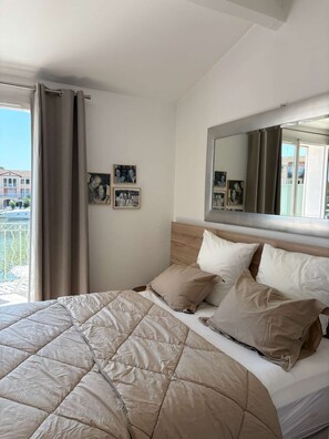 Ein Hotelzimmer mit Bett, großem Fenster mit Vorhängen, wandmontiertem Spiegel und gerahmten Bildern.