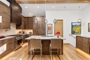 A modern kitchen with wooden cabinets, a central island with bar stools, pendant lighting, and a framed landscape painting on the wall. - Newly Listed! Designer Mountain Retreat Steps from the Gondola. Great Winter Availability! (Aspen)