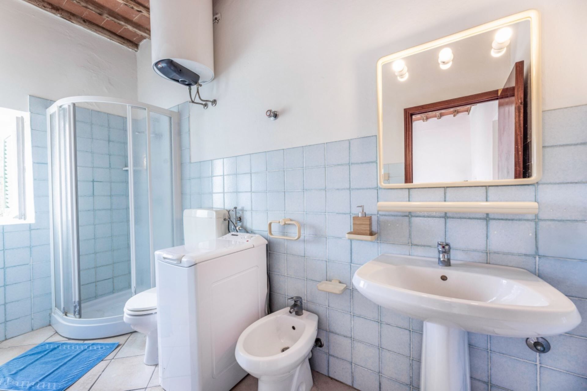 Et badeværelse med bruser, toilet, bidet, vask og et spejl med lys.