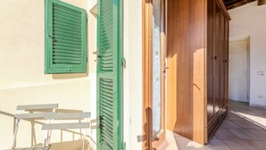 Een kamer met een houten deur, een raam met groene luiken en een kleine tafel.