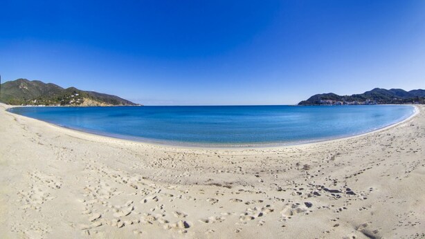 Een breed zandstrand met helderblauw water en bergen op de achtergrond.