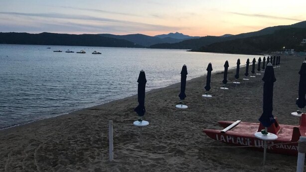 Een strand met rijen parasols en ligbedden, een redderspost en boten op het water.