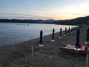 Una spiaggia con file di ombrelloni e lettini, una postazione di salvataggio e barche sull'acqua.
