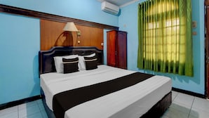 A bedroom with a bed, a wooden headboard, a nightstand, a lamp, a wardrobe, and a window with curtains. - OYO 95140 Metro Guest House (Yogyakarta)