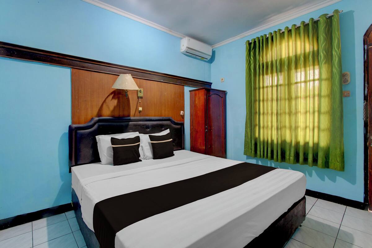 A bedroom with a bed, a wooden headboard, a nightstand, a lamp, a wardrobe, and a window with curtains.