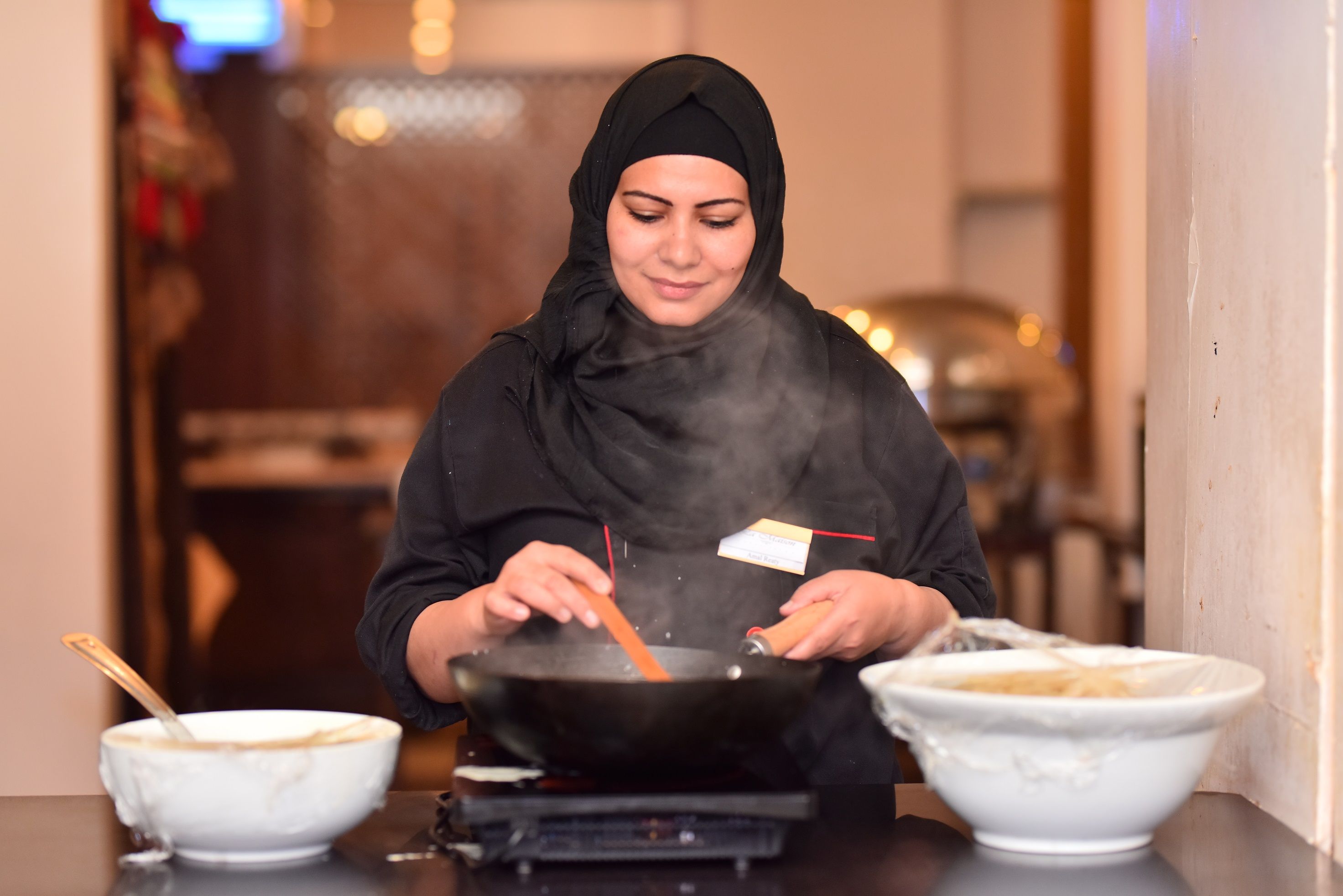 A person cooking in a kitchen with a wok on a portable stove, two bowls on the counter, and a blurred background.