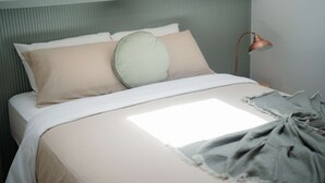 En pænt redt seng med et lyst sengetæppe og puder, en lampe ved siden af og et vindue der lukker naturligt lys ind.