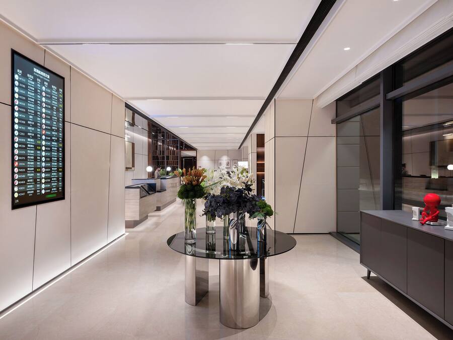 A modern hotel lobby with a digital directory, a round glass table with floral arrangements, and a minimalist reception desk.