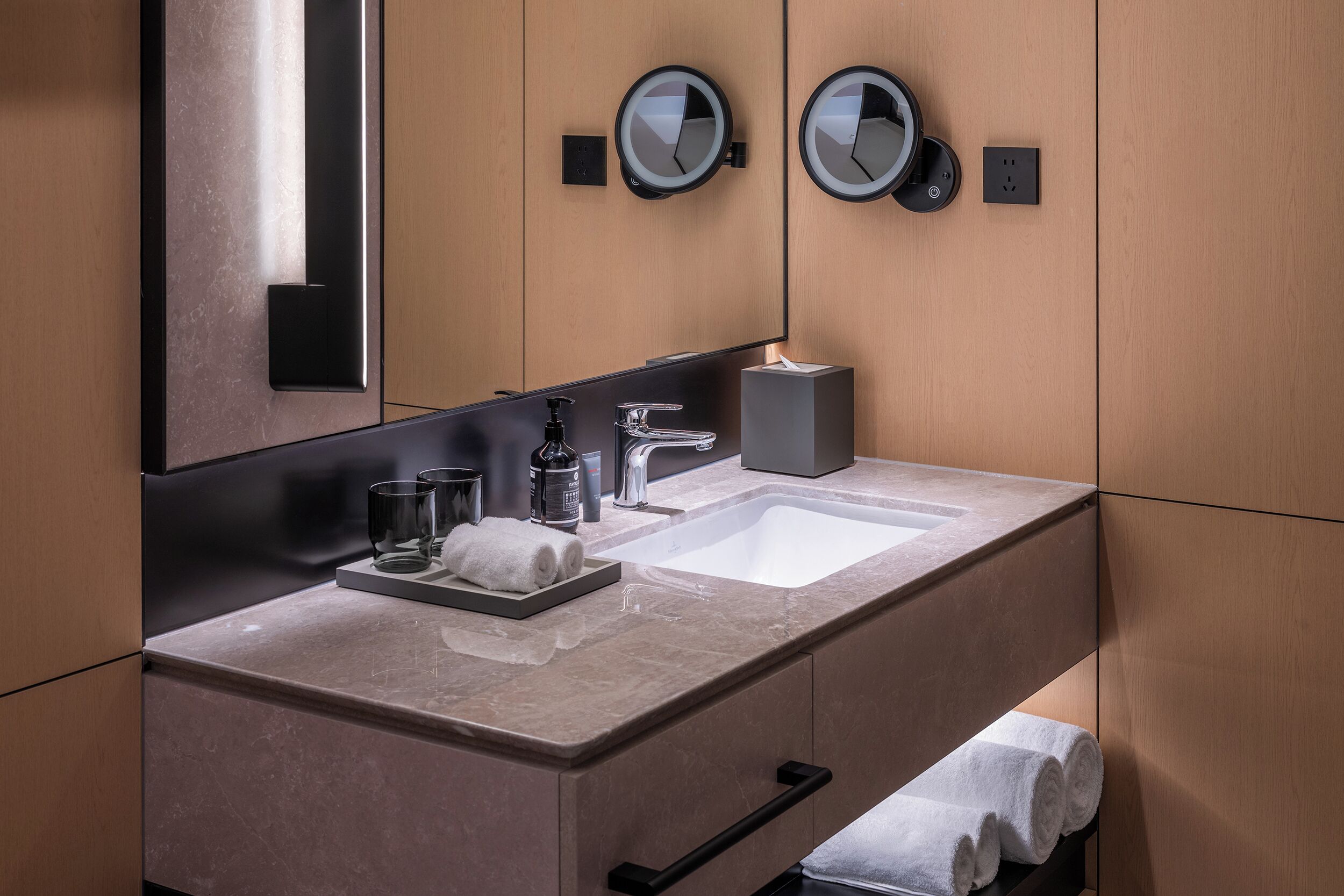 A modern bathroom with a marble sink, two round mirrors, a towel rack, and a soap dispenser.