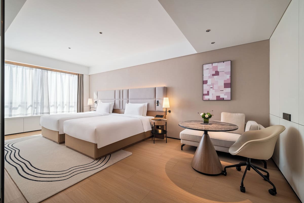 A modern hotel room with two beds, a sofa, a small table, and a large window.