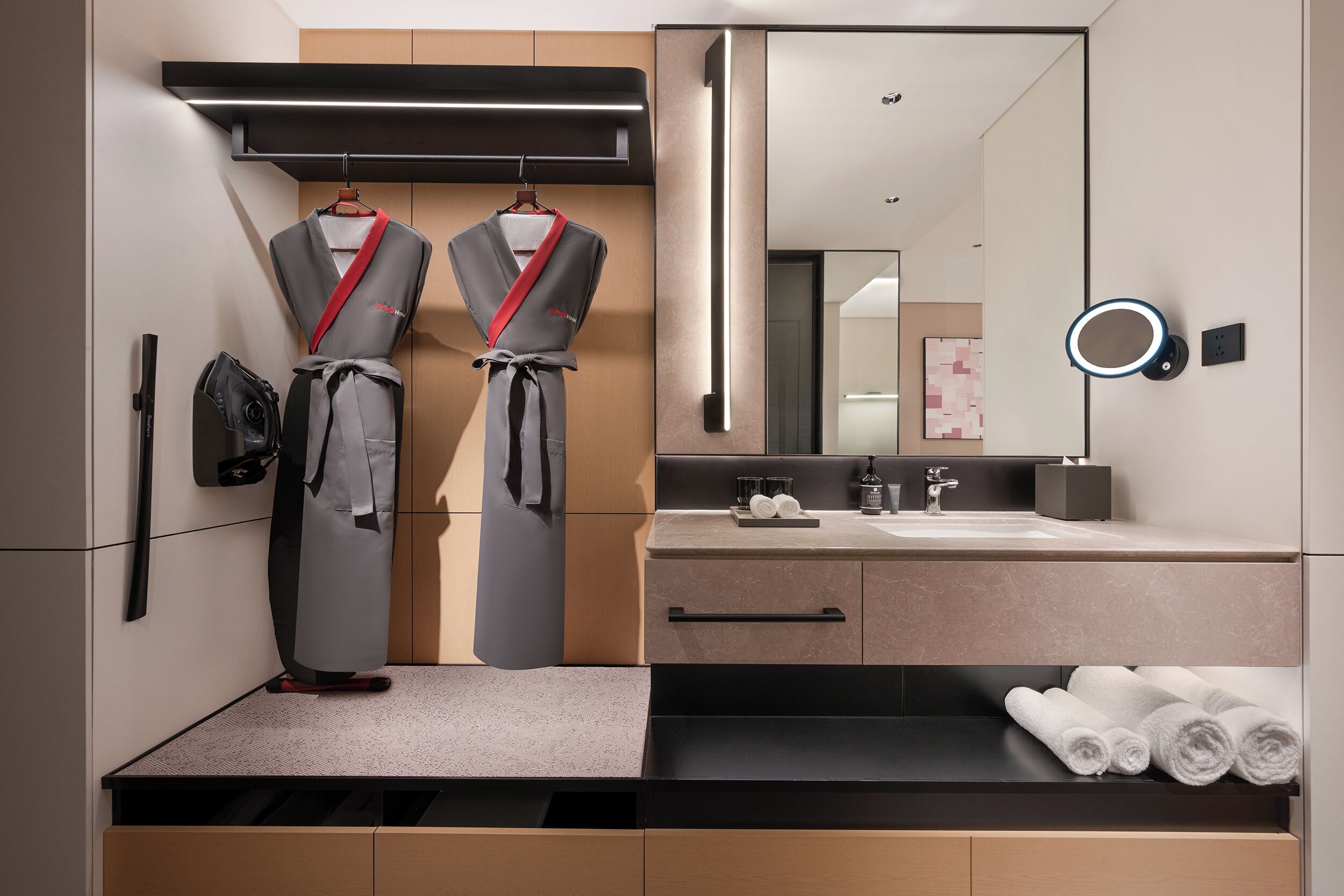 A modern bathroom with a large mirror, a towel rack with robes, a sink with a countertop, and a towel shelf.
