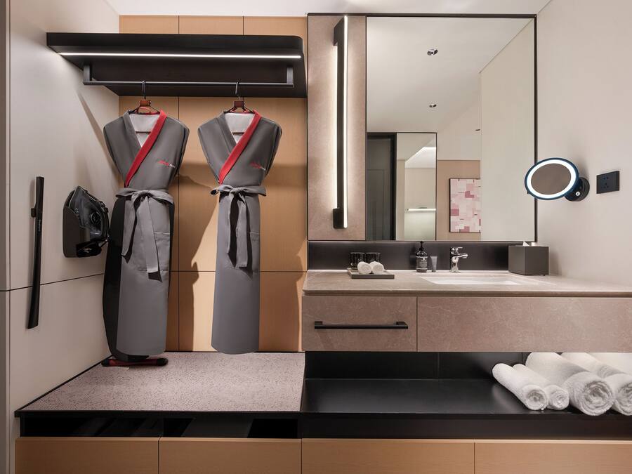A modern bathroom with a large mirror, a towel rack with robes, a sink with a countertop, and a towel shelf.