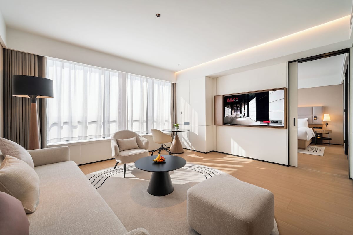A modern hotel room with a sofa, armchair, round coffee table, and a TV in the background.