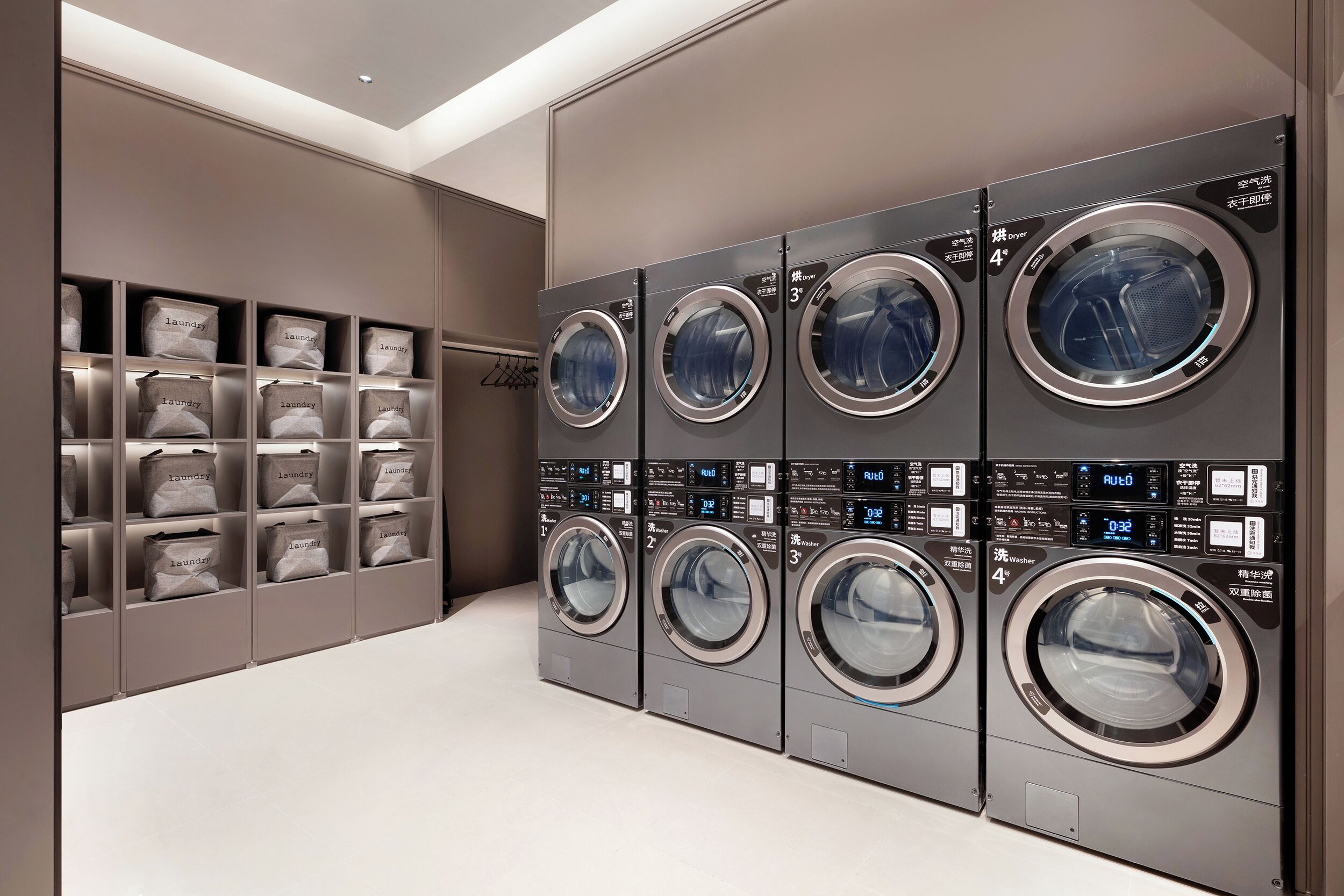 A row of four front-loading washing machines with digital displays, arranged in a modern laundry room with storage shelves.